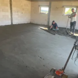 Wykonawca w Kamionce wyrównuje świeżo wylaną posadzkę jastrychową w surowym wnętrzu, widoczne okna i narzędzia pracy.