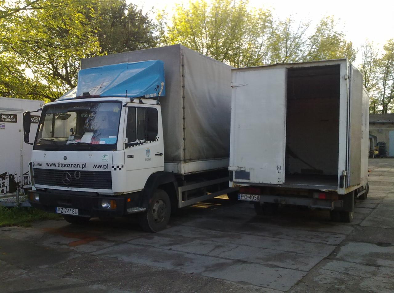 Biała ciężarówka Mercedes z otwartą naczepą na betonowym placu, widoczne tablice rejestracyjne PZ-741BU i PO-4058, drzewa w tle, niebieska plandeka na kabinie.