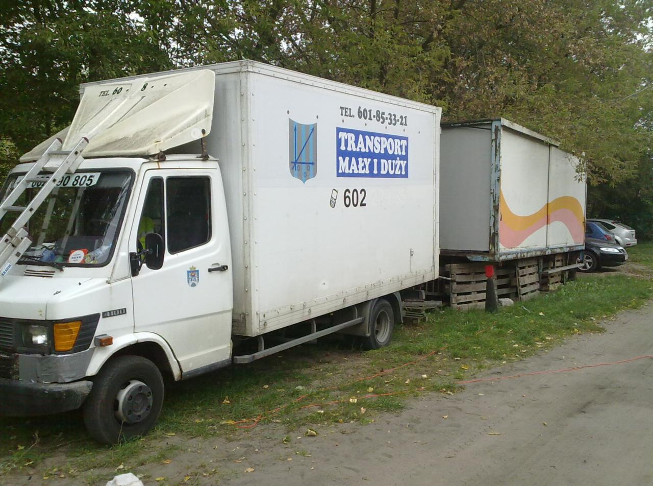 Biała ciężarówka z plandeką i drabiną, z napisem 'Transport mały i duży' oraz numerem telefonu, zaparkowana obok przyczepy na tle drzew.