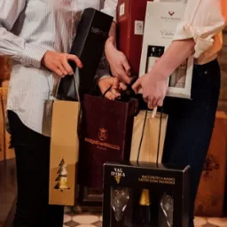 Dwie osoby trzymają w rękach zestawy prezentowe z alkoholem, w tym butelki w ozdobnych pudełkach i torbach prezentowych. Widoczne marki to Villa Maria, Marques de Murrieta i Val D'Oca. W tle półki...