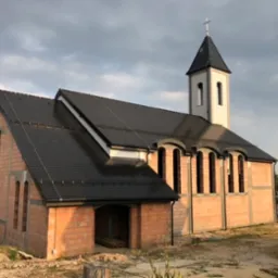 Kościół w trakcie budowy: surowe mury z cegły, czarny dach z dachówki, biała wieża z krzyżem na szczycie, widoczne otwory okienne.