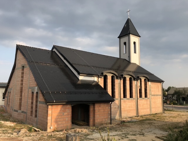 Kościół w trakcie budowy: surowe mury z cegły, czarny dach z dachówki, biała wieża z krzyżem na szczycie, widoczne otwory okienne.