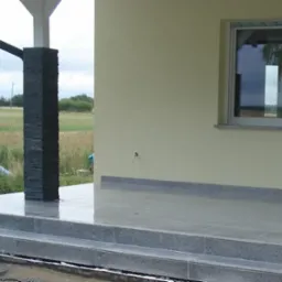 Wykończony taras domu z szarych płytek i kamiennymi słupkami. Elewacja kremowa, okno z szarą ramą. Widok na zielone pola w tle.