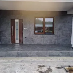 Wykończony taras z szarymi, wielkoformatowymi płytkami imitującymi kamień, z brązowymi drzwiami wejściowymi i oknem, elewacja częściowo wykończona płytkami, widoczny fragment kostki brukowej przed...