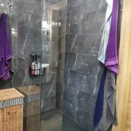 Nowoczesna kabina prysznicowa z ciemnymi płytkami imitującymi marmur, widoczny kosz na pranie z wikliny i zestaw kosmetyk&oacute;w wewnątrz kabiny.