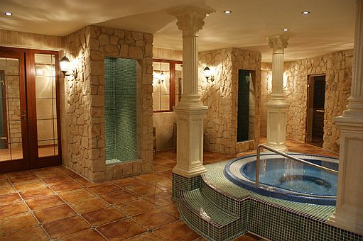 Wnętrze luksusowej łazienki z jacuzzi, kamiennymi ścianami, kolumnami i mozaiką, oświetlone ciepłym światłem.