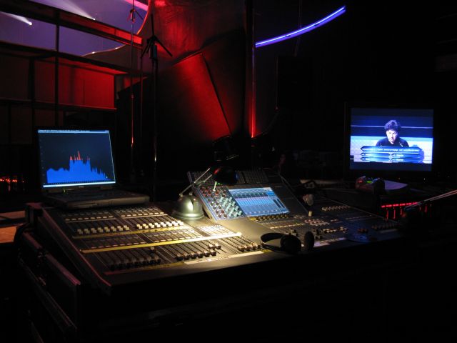 Stanowisko mikserskie z laptopem wyświetlającym analizator dźwięku i monitorem z widocznym fragmentem teleturnieju, oświetlone czerwonym i niebieskim światłem.