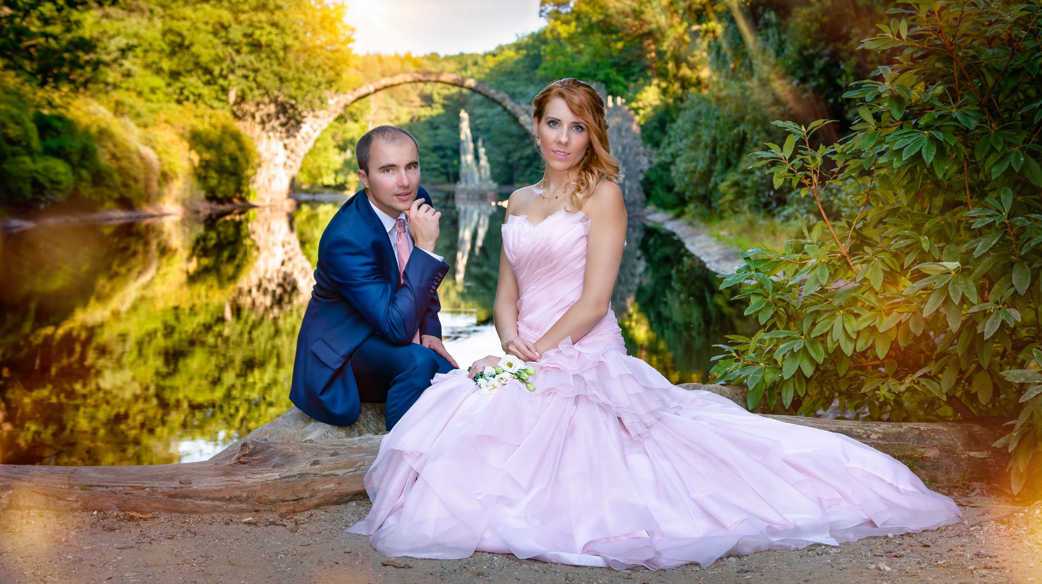 Para młoda siedzi na pniu drzewa nad wodą z widokiem na kamienny most w parku Kromlau, kobieta w różowej sukni ślubnej trzyma bukiet.