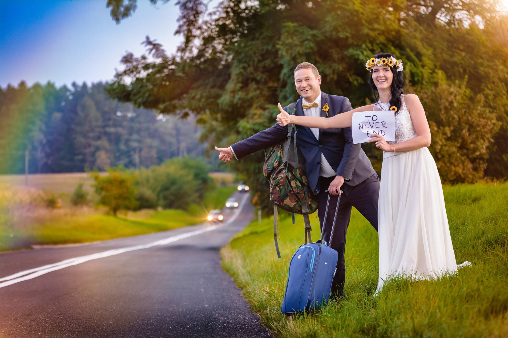 Para młoda w podróży poślubnej autostopem: pan młody w garniturze z plecakiem i walizką, panna młoda w sukni ślubnej z wiankiem słoneczników trzyma kartkę z napisem 'To Never End'.