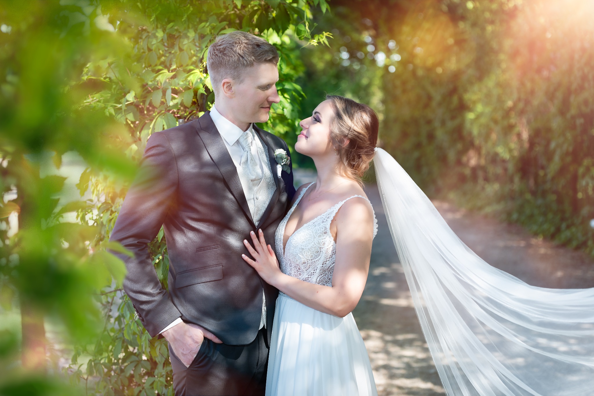 Młoda para w strojach ślubnych, spoglądająca na siebie z czułością w otoczeniu zieleni, z promieniami słońca przebijającymi przez liście.