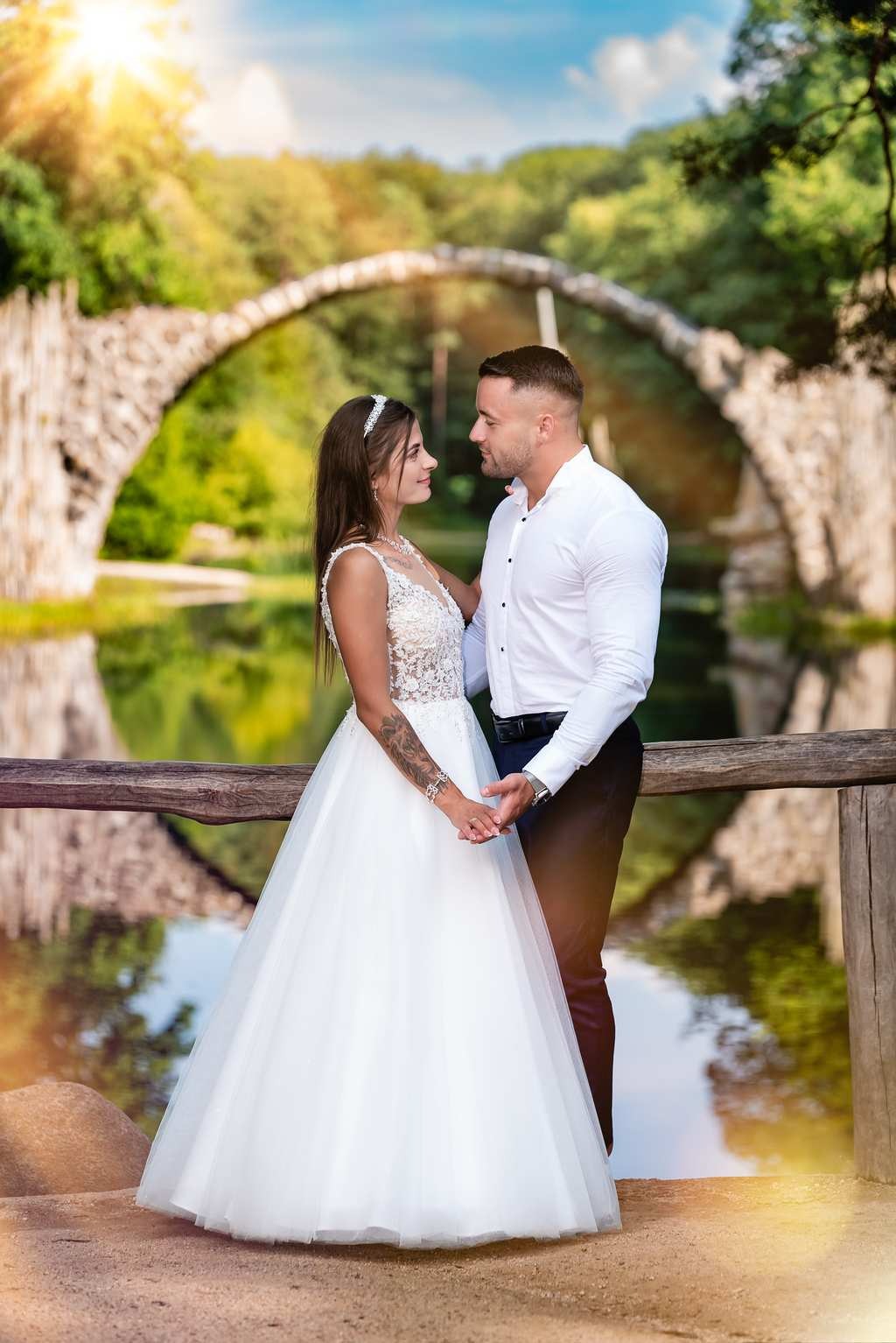 Para młoda trzymająca się za ręce na drewnianym moście z widokiem na Rakotzbrücke, Panna Młoda ma tatuaż na ramieniu, delikatna suknia ślubna z koronkową górą i tiarą.