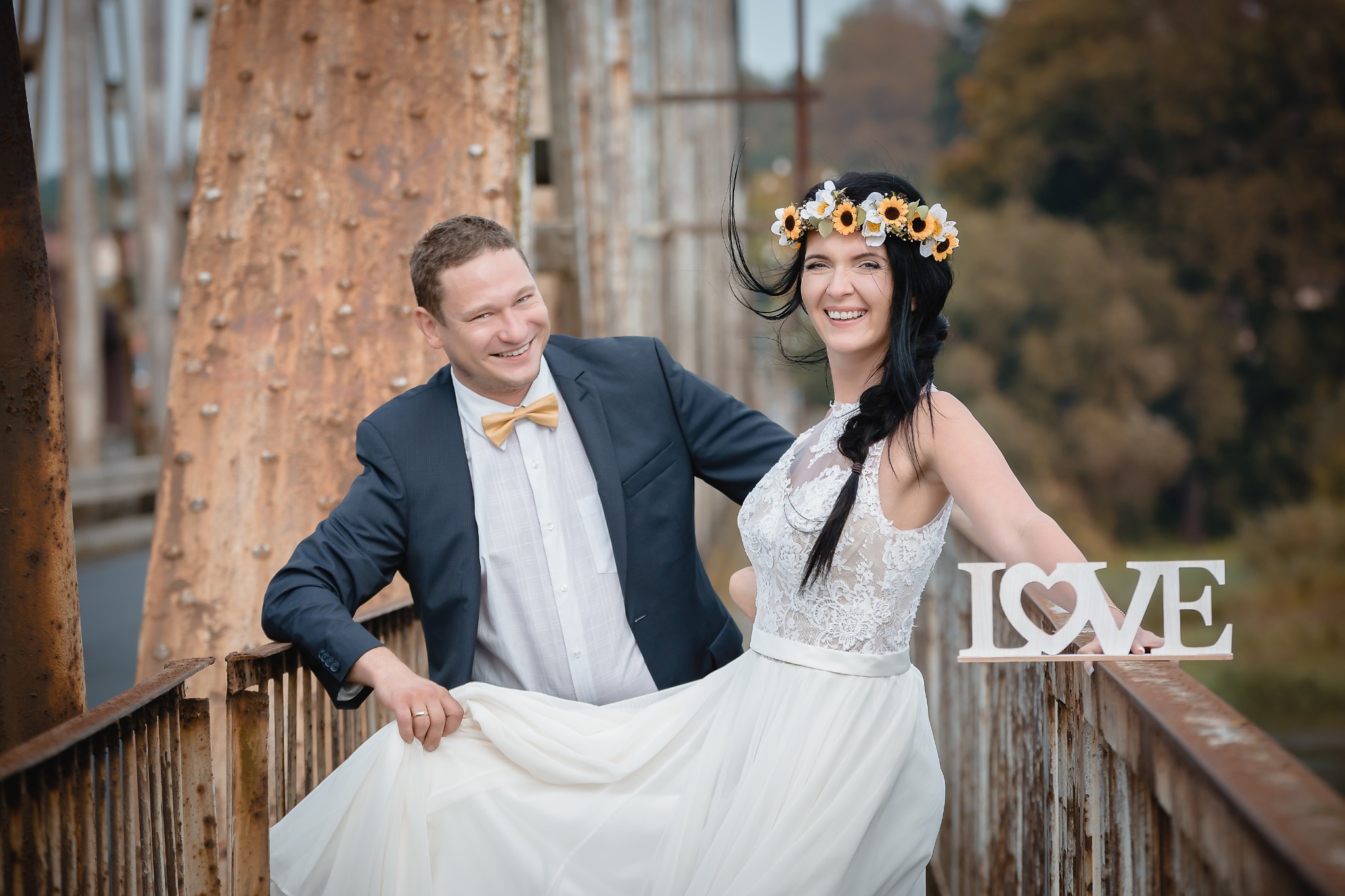 Uśmiechnięta para młoda w strojach ślubnych pozuje na tle zardzewiałego mostu, panna młoda trzyma drewniany napis LOVE.