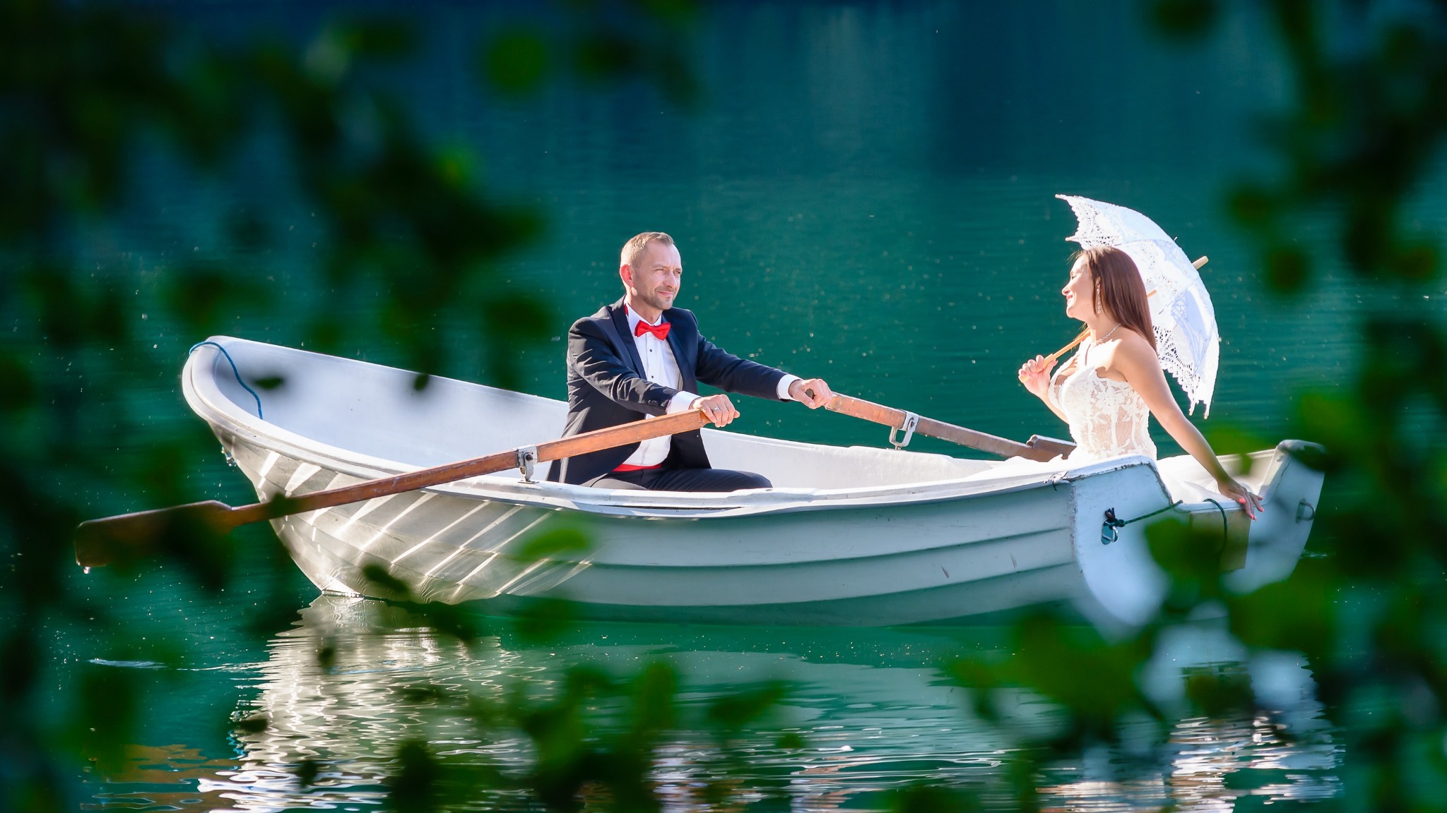 Para młoda w łódce na jeziorze, mężczyzna w smokingu wiosłuje, kobieta w białej sukni trzyma koronkowy parasol, romantyczna sceneria z zielonym tłem.