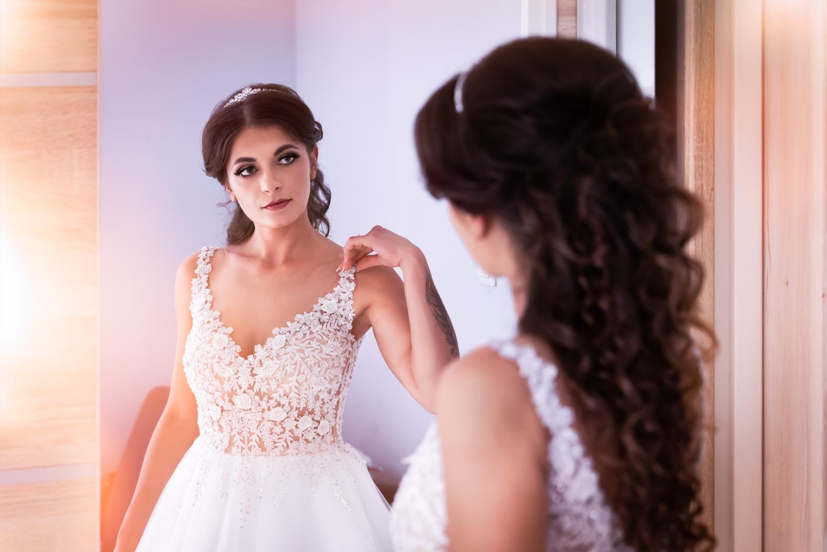 Panna młoda w koronkowej sukni ślubnej, spoglądająca w lustro, z widocznym tatuażem na ramieniu.