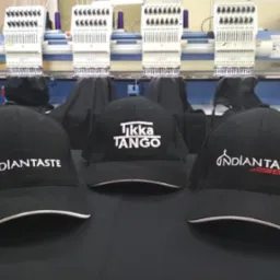 Trzy czarne czapki z daszkiem z różnymi białymi napisami, ustawione na tle maszyn hafciarskich.