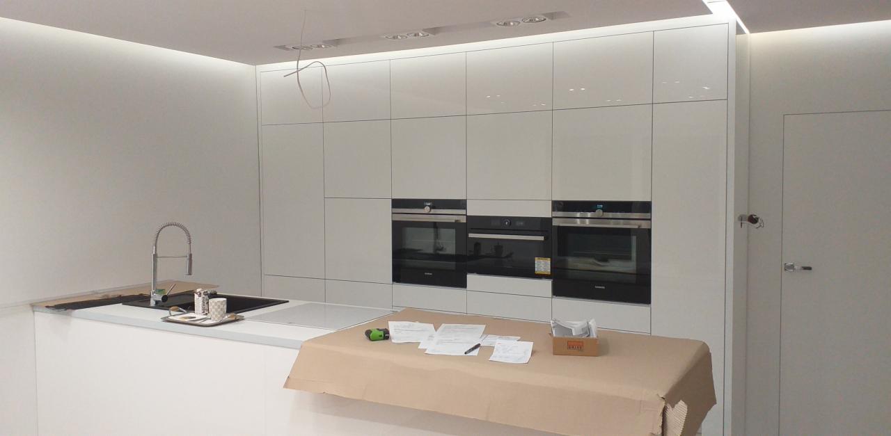 Nowoczesna, biała kuchnia z zabudową pod sufit, trzema piekarnikami, wyspą z umywalką i płytą indukcyjną, blat wyspy przykryty folią ochronną, widoczne dokumenty i narzędzia.