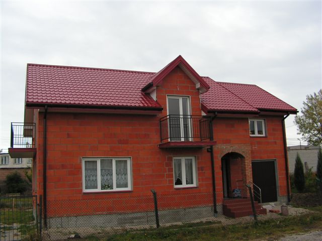 Nowy dom w stanie surowym otwartym z czerwoną, błyszczącą dachówką, widoczne rynny i obróbki blacharskie.