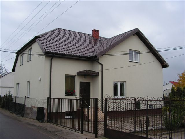 Dwupiętrowy dom jednorodzinny z brązowym dachem z blachodachówki, jasną elewacją i brązowymi rynnami, otoczony metalowym ogrodzeniem, widoczny z poziomu ulicy w pochmurny dzień.