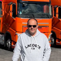 Mężczyzna w bluzie Lacoste stoi obok pomarańczowej ciężarówki Volvo F11 i beczki z napisem Imperium Truck