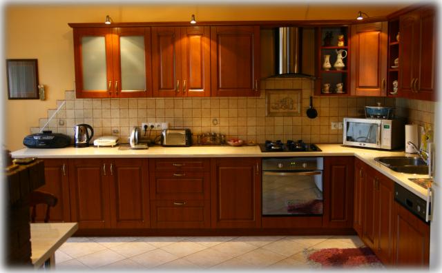 Kompletna zabudowa kuchenna z drewnianymi frontami w odcieniu ciepłego brązu, z blatem w jasnym kolorze, wyposażona w sprzęt AGD, widoczna z perspektywy szerokokątnej.