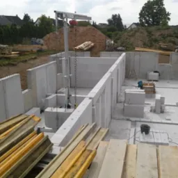 Fundamenty domu z betonu komórkowego. Widoczny dźwig, drewno, bloczki i ziemia w tle. Ujęcie z góry ukazuje postęp prac.