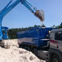 Niebieska koparka Komatsu PC210NLC załadowuje piasek do niebieskiej naczepy ciężarówki MAN z logo firmy Slota Industry S.C. na tle lasu i błękitnego nieba.