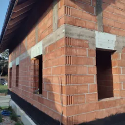 Wznoszenie ścian zewnętrznych domu z czerwonej cegły ceramicznej, widoczne otwory okienne i warstwa izolacji przeciwwilgociowej fundamentów. Na drugim planie stos cegieł i częściowo zadaszona...
