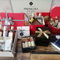 Zestaw eleganckich, kolorowych pudełek prezentowych z alkoholami i słodyczami, z logo 'Piwniczka Smaku' w tle, na drewnianym blacie.