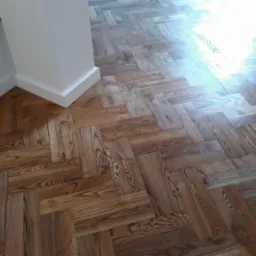 Ułożona parkietowa podłoga w jodełkę, z widocznym fragmentem ściany i listwy przypodłogowej, oświetlona naturalnym światłem.