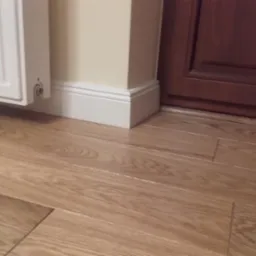 Ułożone panele podłogowe w odcieniach dębu, widoczne łączenia, białe listwy przypodłogowe przy ścianie pomalowanej na kremowo, fragment brązowych drzwi.