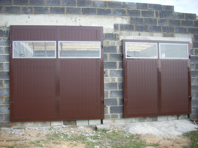 Brama garażowa dwuskrzydłowa, brązowa, z dwoma przeszkleniami na skrzydło, zamontowana w surowym murze z bloczków betonowych.