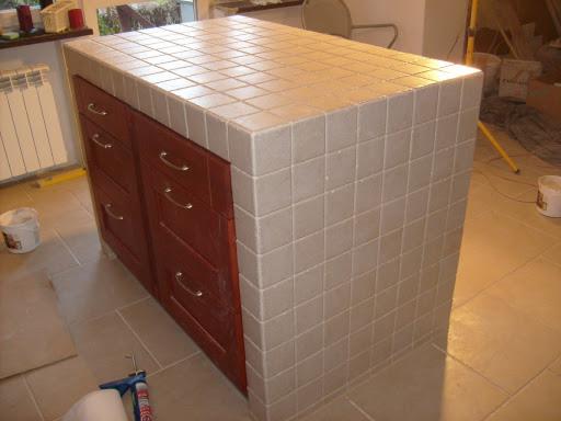 Wyspa kuchenna z ciemnymi drewnianymi szafkami i blatem wykończonym jasnymi płytkami ceramicznymi, widoczne narzędzia i materiały budowlane na podłodze.