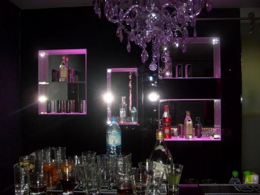 Lustrzane półki z fioletowym podświetleniem, na których stoją butelki z alkoholem, nad nimi wisi kryształowy żyrandol, a na blacie stoją szklanki i butelki.
