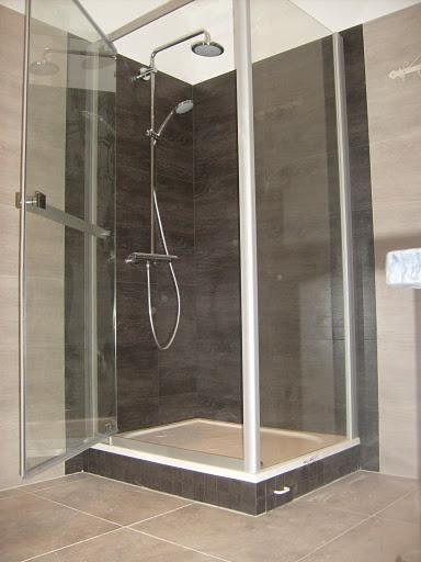 Szklana kabina prysznicowa z deszczownicą i baterią termostatyczną, wykończona płytkami w odcieniach szarości, z częściowo otwartymi drzwiami.