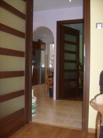 Widok na kuchnię przez otwarte, wewnętrzne drzwi z drewnianymi ramami i przeszkleniami, podłoga wyłożona płytkami ceramicznymi.