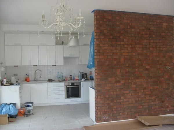 Wnętrze kuchni w trakcie remontu, widoczne białe szafki kuchenne, ozdobny żyrandol i ściana z cegły w odcieniach czerwieni i brązu, zabezpieczona niebieską folią malarską.