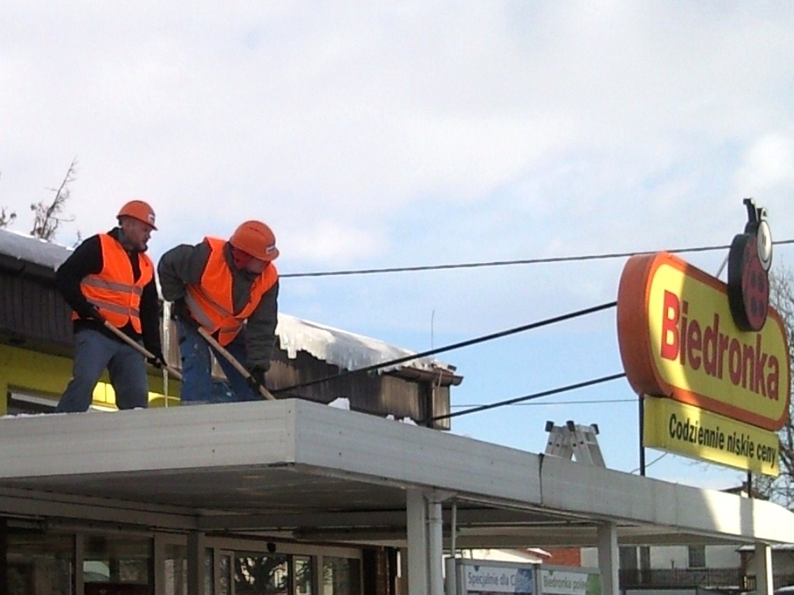 Dwóch pracowników w pomarańczowych kamizelkach i kaskach odśnieża dach pawilonu handlowego Biedronka za pomocą łopat.