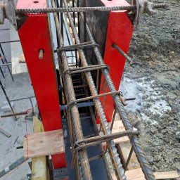 Przemysław Pytel - Zbrojenie pionowe w czerwonym szalunku, widoczne pręty i strzemiona. Konstrukcja wsparta na drewnianych belkach, w tle ziemia. Detal budowy.