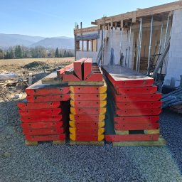 Przemysław Pytel - Stos czerwonych szalunków na żwirowym podłożu, w tle budowa domu z białych bloczków i widok na góry w słoneczny dzień. Widoczne elementy konstrukcji i podpory.