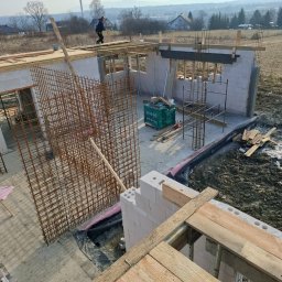Przemysław Pytel - Budowa domu z betonu komórkowego: widok z góry na wznoszone ściany, zbrojenie i szalunki. W tle krajobraz z zabudowaniami i kominem.