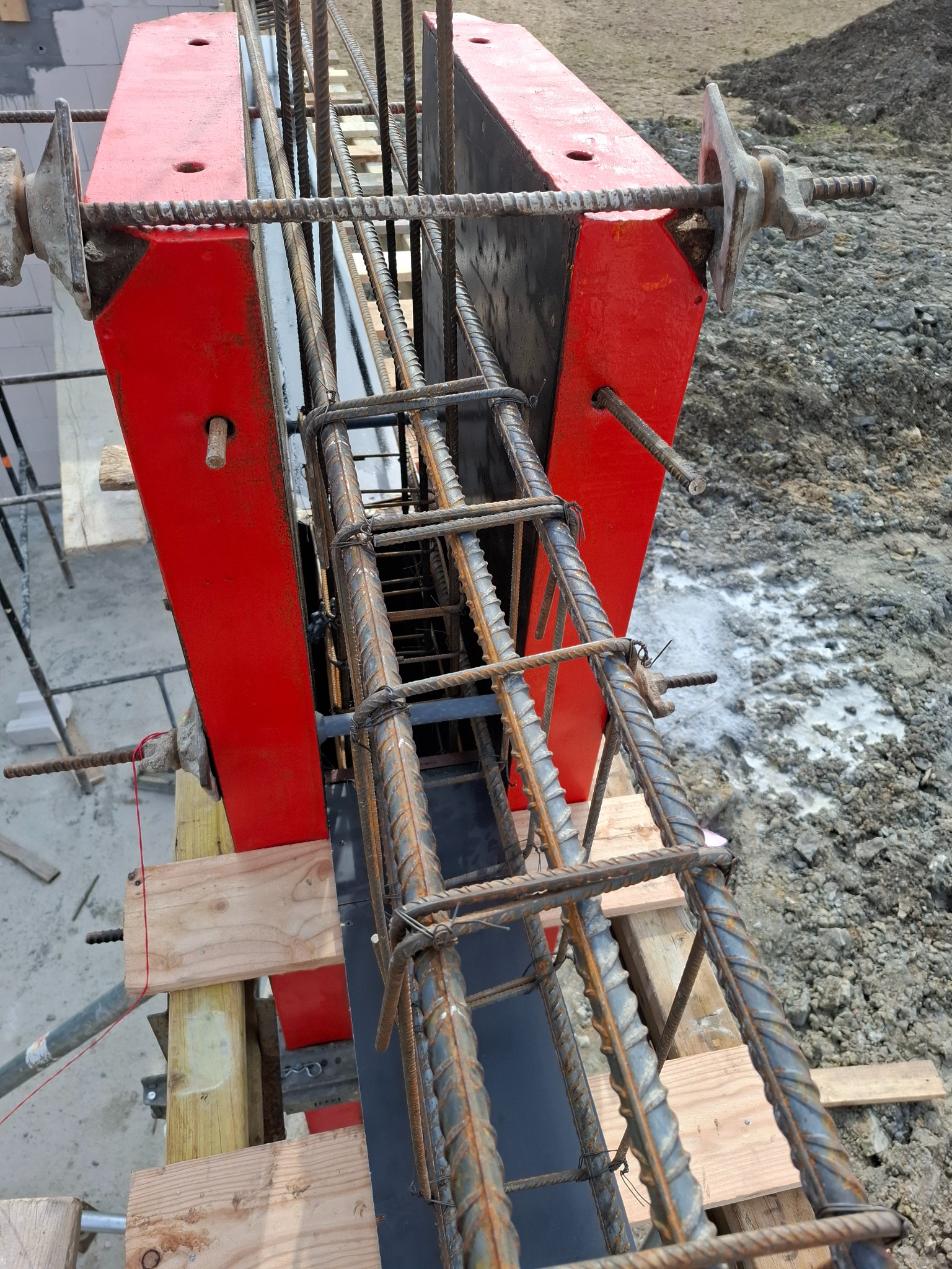 Zbrojenie pionowe w czerwonym szalunku, widoczne pręty i strzemiona. Konstrukcja wsparta na drewnianych belkach, w tle ziemia. Detal budowy.