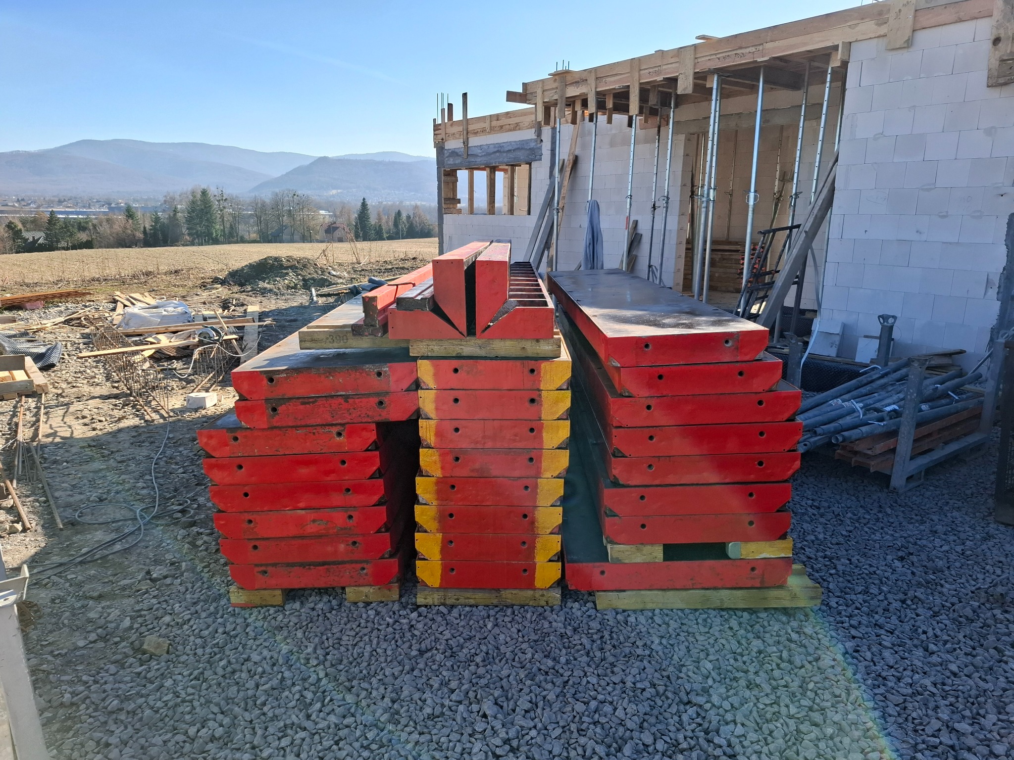 Stos czerwonych szalunków na żwirowym podłożu, w tle budowa domu z białych bloczków i widok na góry w słoneczny dzień. Widoczne elementy konstrukcji i podpory.