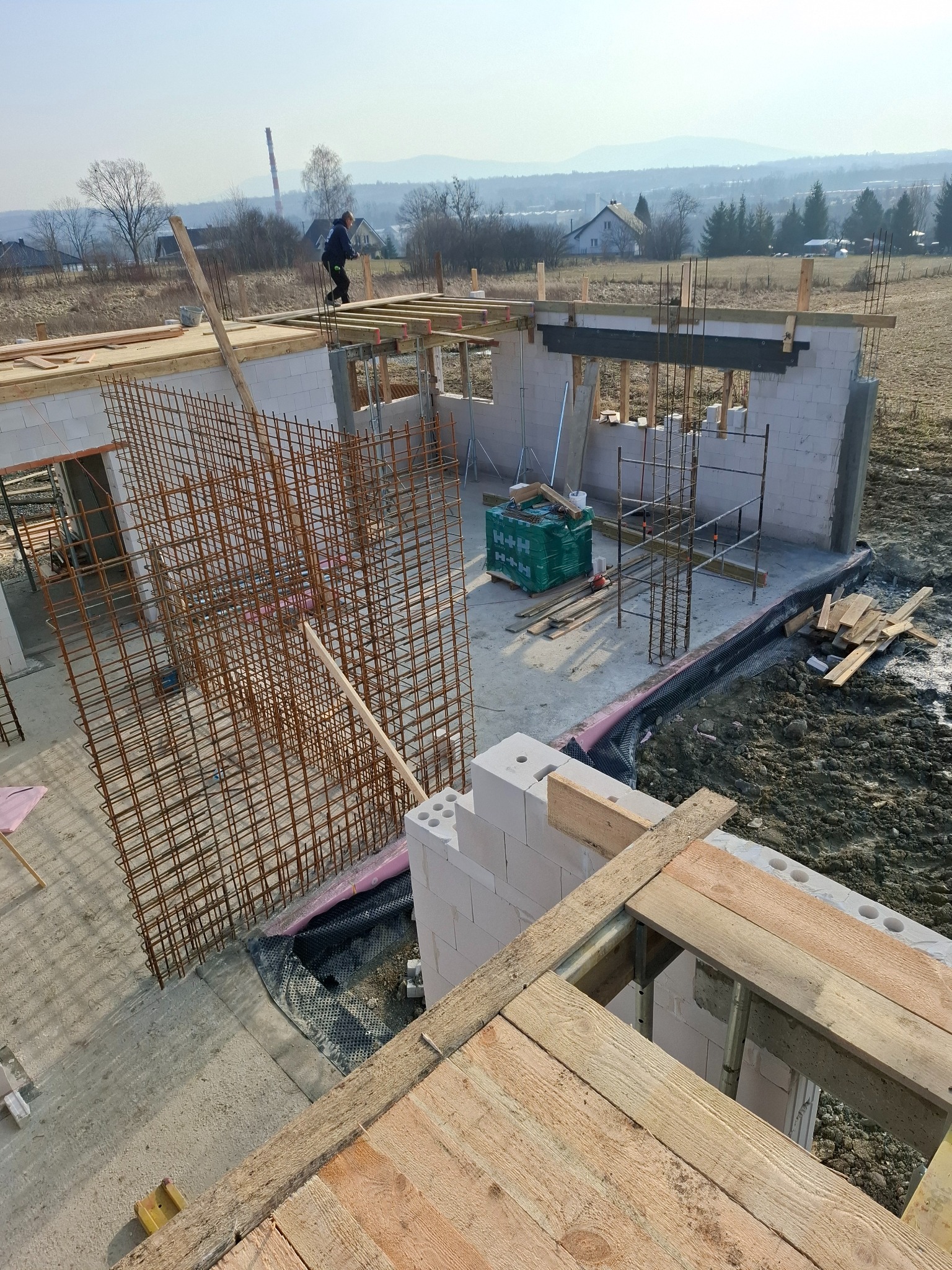 Budowa domu z betonu komórkowego: widok z góry na wznoszone ściany, zbrojenie i szalunki. W tle krajobraz z zabudowaniami i kominem.