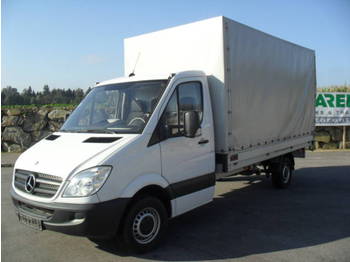 Biały samochód dostawczy Mercedes Sprinter z otwartymi drzwiami kierowcy, zaparkowany na parkingu z zielenią w tle.