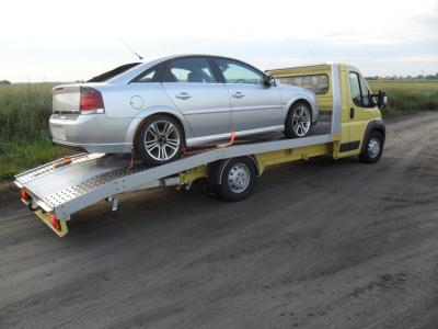 Żółta autolaweta z załadowanym srebrnym samochodem osobowym marki Opel Vectra na platformie, zabezpieczonym pomarańczowymi pasami transportowymi, na polnej drodze.