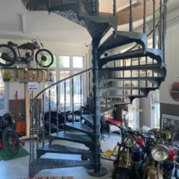 Kuta, spiralna klatka schodowa w warsztacie motocyklowym, otoczona zabytkowymi motocyklami i trofeami.