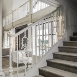 Drewniane schody z ozdobną, białą balustradą i widokiem na salon w tle, oświetlone naturalnym światłem.
