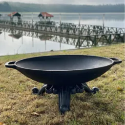 Czarny, metalowy palenisko ogrodowe w kształcie misy na ozdobnej podstawie, ustawione na trawie, z widokiem na jezioro i pomost w tle, pochmurny dzień.