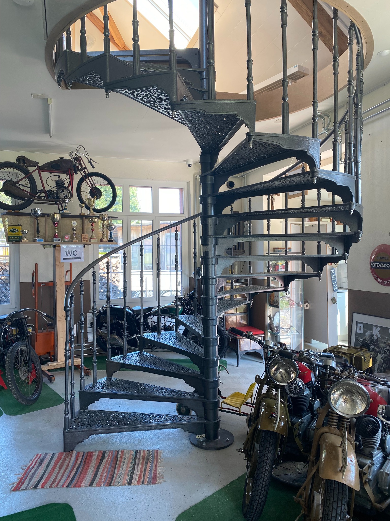Kuta, spiralna klatka schodowa w warsztacie motocyklowym, otoczona zabytkowymi motocyklami i trofeami.