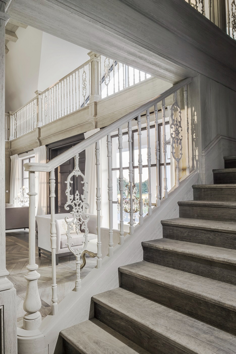 Drewniane schody z ozdobną, białą balustradą i widokiem na salon w tle, oświetlone naturalnym światłem.